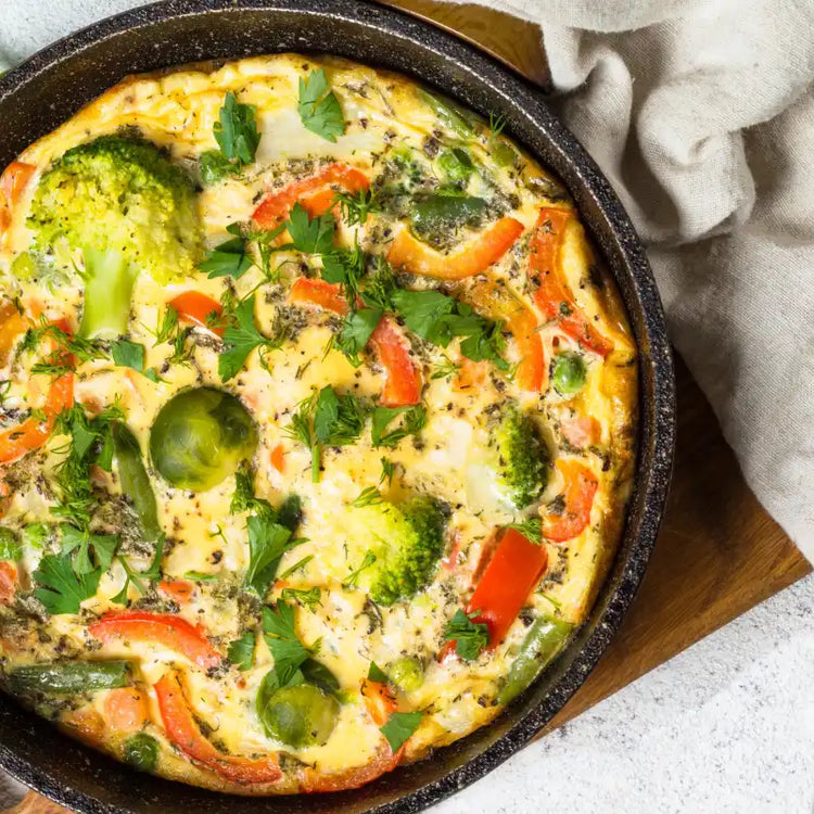 Colorful vegetable frittata in a black skillet, filled with broccoli, Brussels sprouts, red peppers, and herbs.