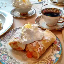 Delicate German-style crêpes filled with cream and fruit, dusted with powdered sugar and topped with whipped cream, served on fine china with a cup of black coffee.