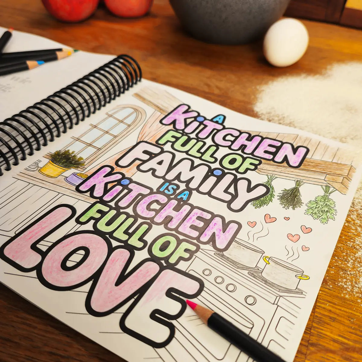 Coloring page from More Baking Just Like Oma Journal Edition, spiral German baking cookbook featuring “A Kitchen Full of Family is a Kitchen Full of Love” design on a wooden table with pencils and flour.