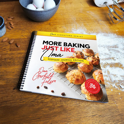 More Baking Just Like Oma Journal Edition spiral cookbook on kitchen table with flour and baking tools, German baking book for home bakers and families.