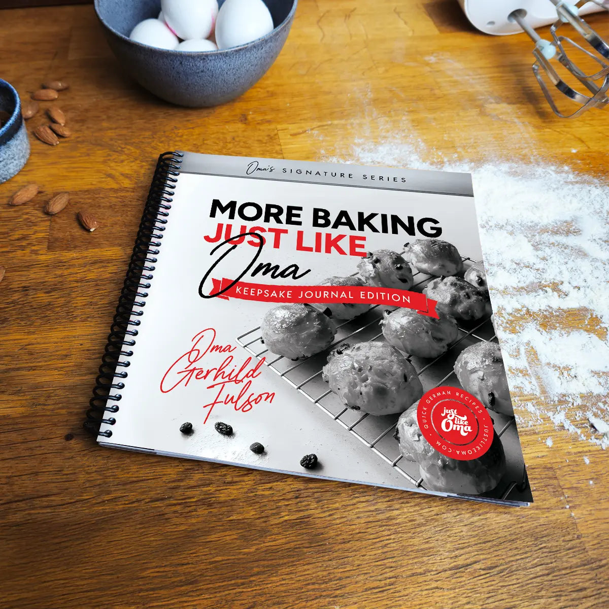 More Baking Just Like Oma Journal Edition black and white spiral cookbook on wooden kitchen table with flour and baking tools, German baking book gift idea.