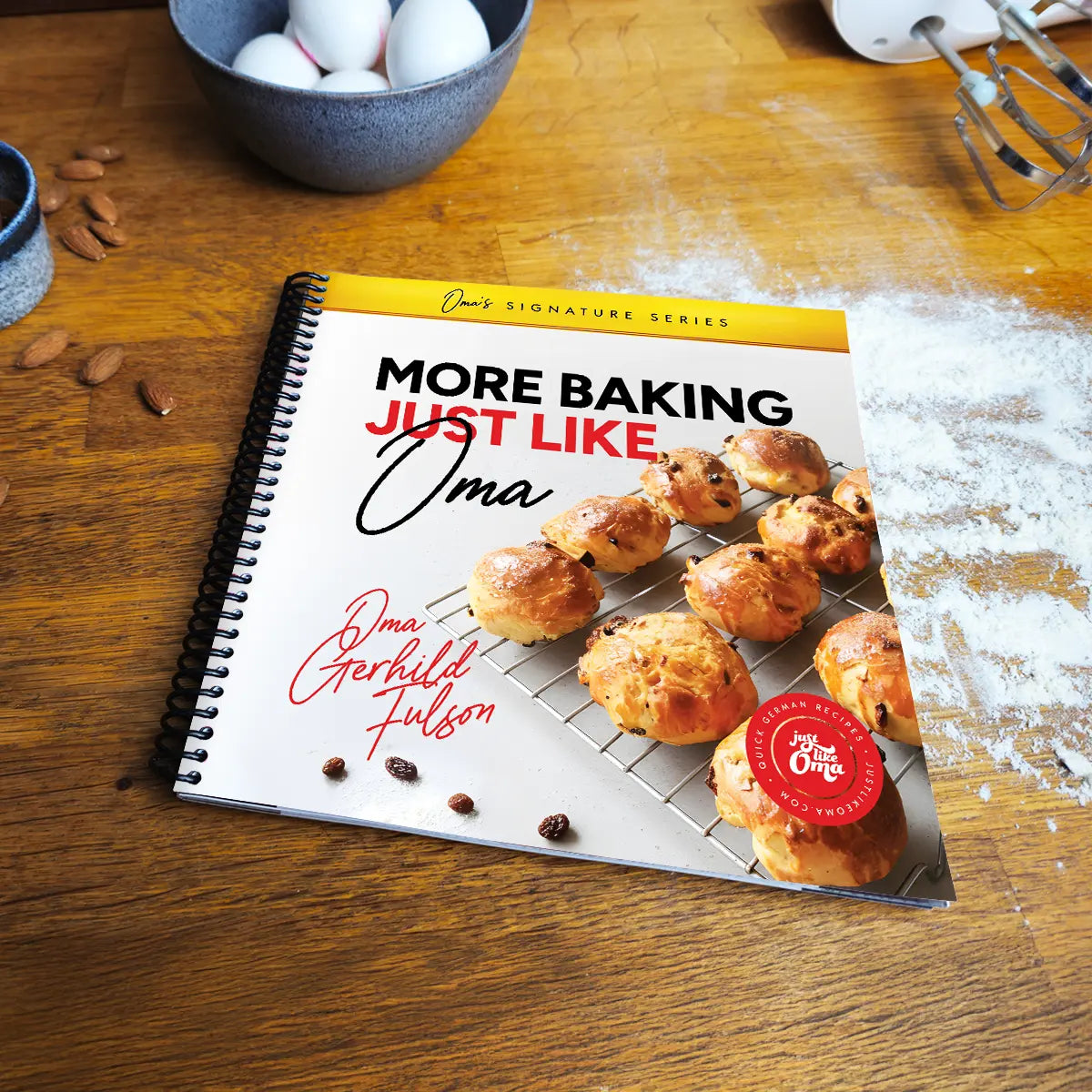 More Baking Just Like Oma spiral cookbook on wooden kitchen table with flour and eggs, authentic German baking book for home bakers and families.