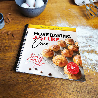 More Baking Just Like Oma spiral cookbook on wooden kitchen table with flour and eggs, authentic German baking book for home bakers and families.
